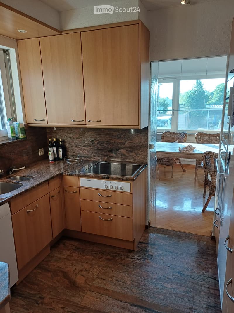 Modern kitchen with light wooden cabinets, marble countertops, and stainless steel appliances. A window offers a view of a dining area with wicker chairs and a table.