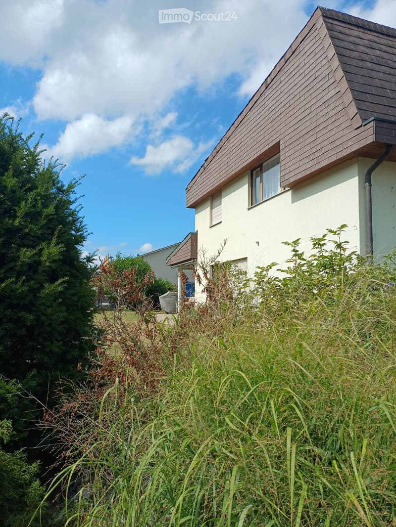 Modern two-story house with white walls and brown wooden roof, surrounded by greenery and tall grass.