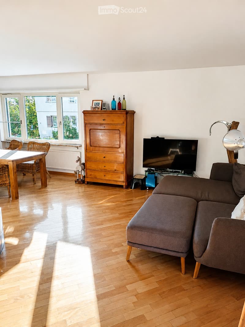 Bright living room with wooden flooring, large windows, a grey sofa, a wooden dining table with chairs, a wooden cabinet, a television, and a small stool.