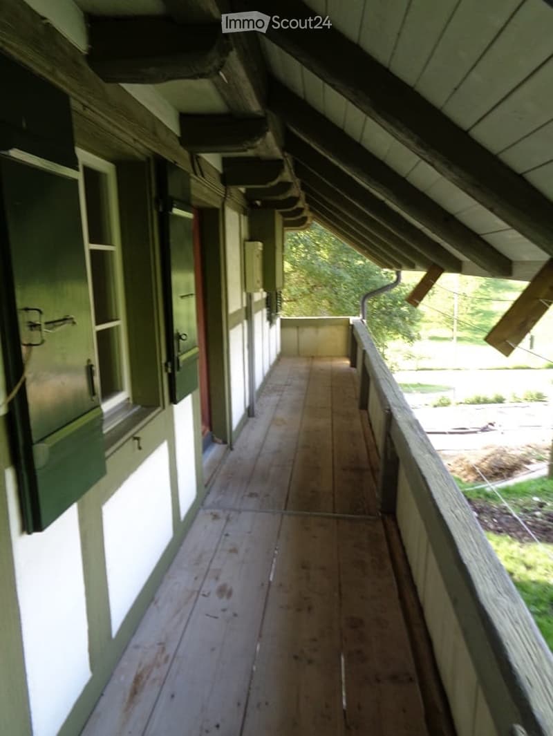 Wooden balcony with slanted roof, green shutters, and a view of greenery outside.
