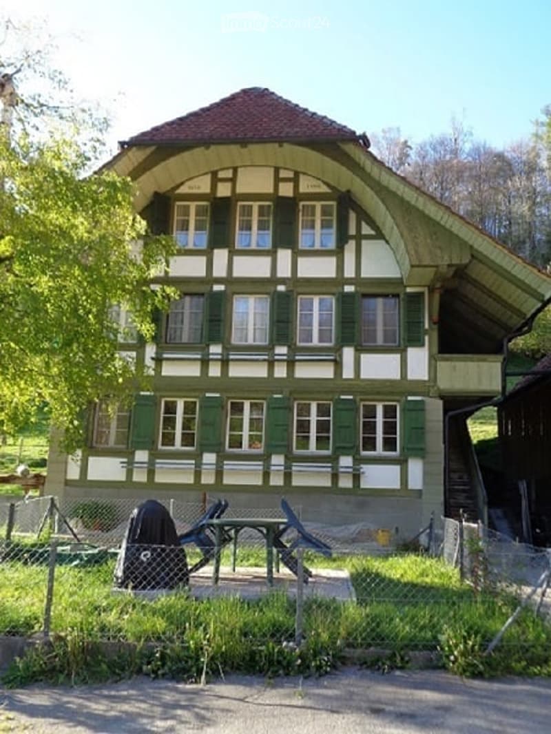 Traditional two-story house with green shutters and white trim, surrounded by a small garden area with a table and chairs.
