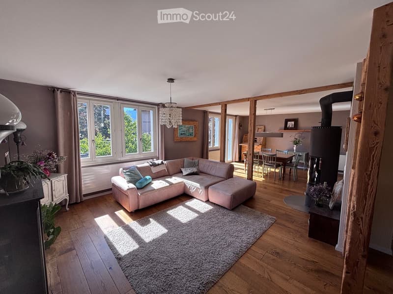 Bright living room with wooden flooring, large windows, a sectional sofa, coffee table, dining area with chairs, wood stove, chandelier, and decorative items.