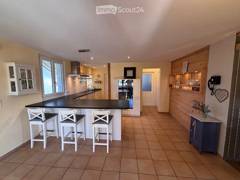 Modern kitchen with a black countertop island, three white bar stools, built-in cabinets, and a small blue cabinet with a flower vase on top. The kitchen features a sink, faucet, refrigerator, oven, and dishwasher, with warm lighting and tiled flooring.