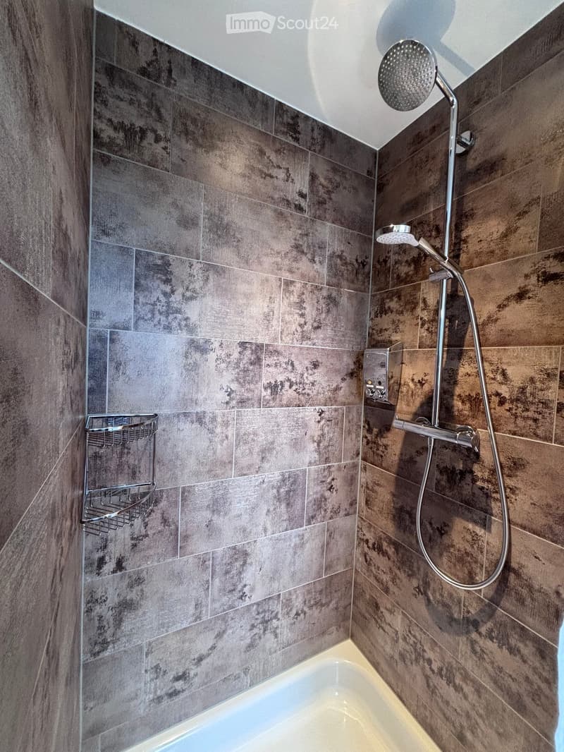 Modern bathroom with a bathtub, a handheld showerhead, a shower hose, a control panel, and a towel rack. The walls are tiled in a dark, textured pattern.