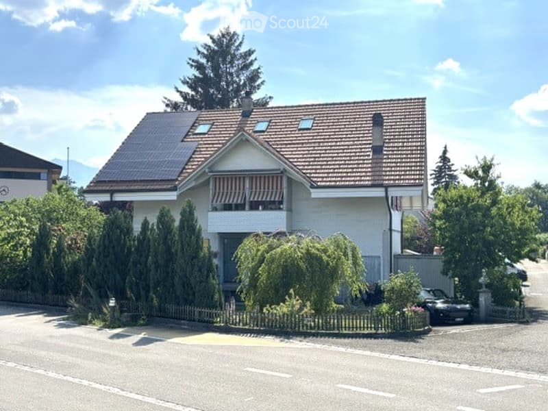Two-story house with a tiled roof, solar panels, white exterior, and a small balcony with striped awnings. Surrounded by greenery and a low fence.