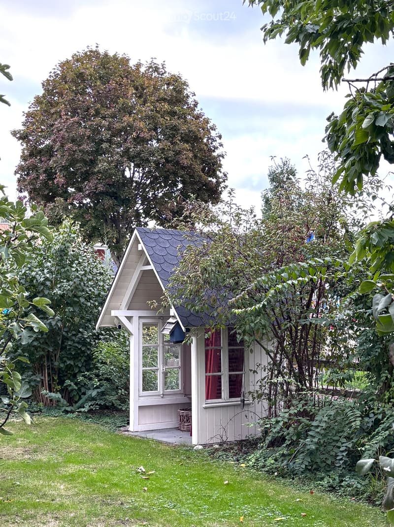A small, white wooden shed with a pointed roof is surrounded by lush greenery and trees in a garden setting.