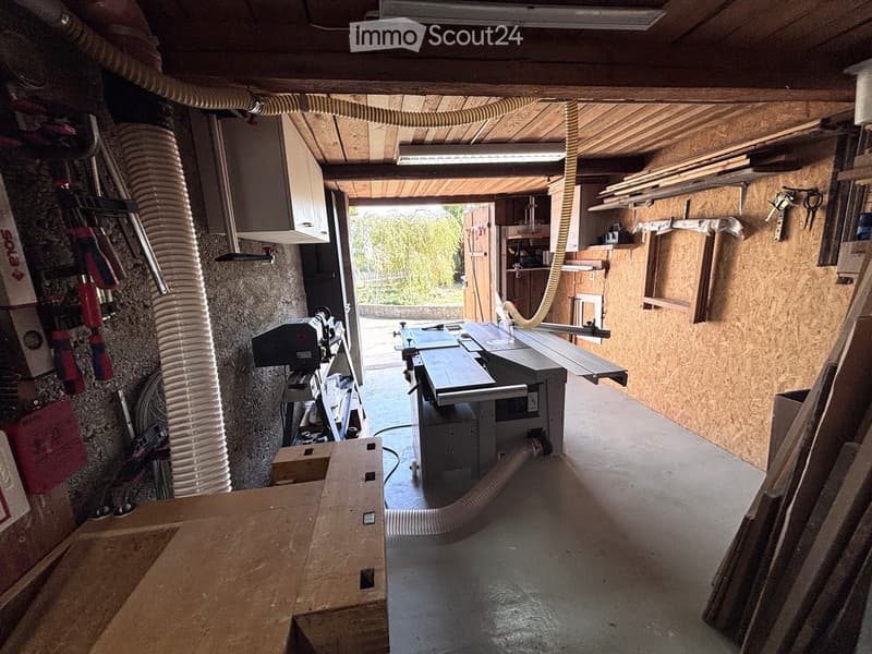 A well-lit workshop with a table saw, workbench, shelves, and cabinets. The space features exposed wooden beams and a concrete floor.