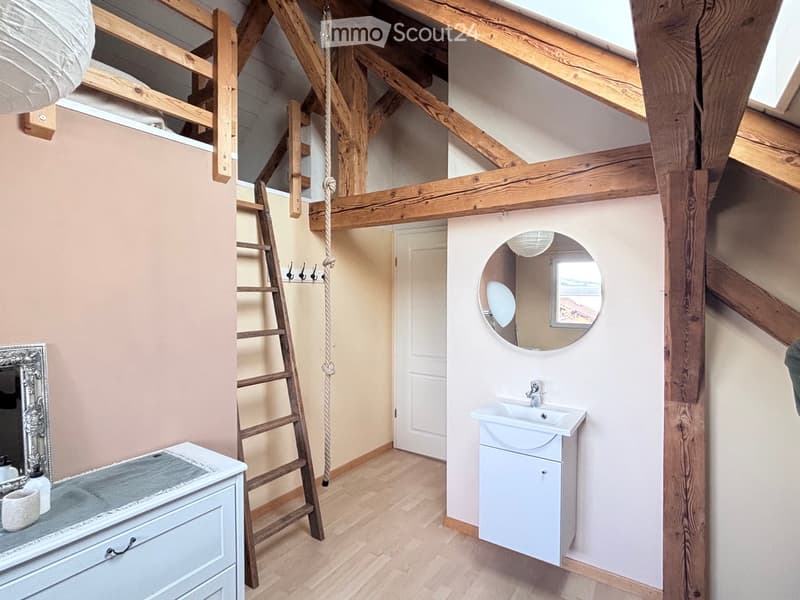 Cozy attic bathroom with exposed wooden beams, a white sink with cabinet, a round mirror, and a wooden ladder leading to a loft area.