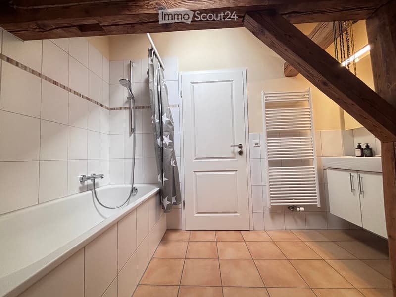 Bathroom with a bathtub, shower, radiator, sink, and cabinet. The walls are tiled, and the floor is tiled with beige tiles.