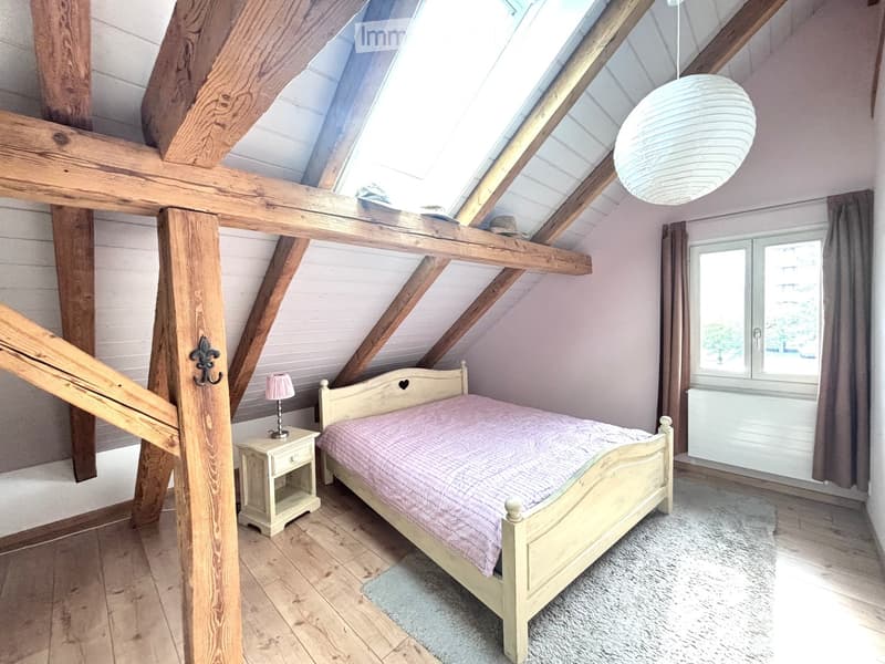 Cozy attic bedroom with wooden beams, skylight, and a single bed with a pink checkered duvet. Features a nightstand with a lamp, wooden floor, and a window with curtains.