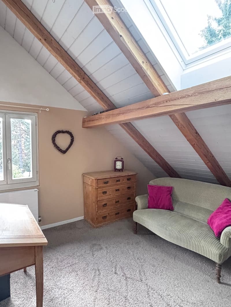 Cozy attic room with exposed wooden beams, a green sofa with pink cushions, a wooden chest of drawers, a small table, and a heart-shaped wall decoration.