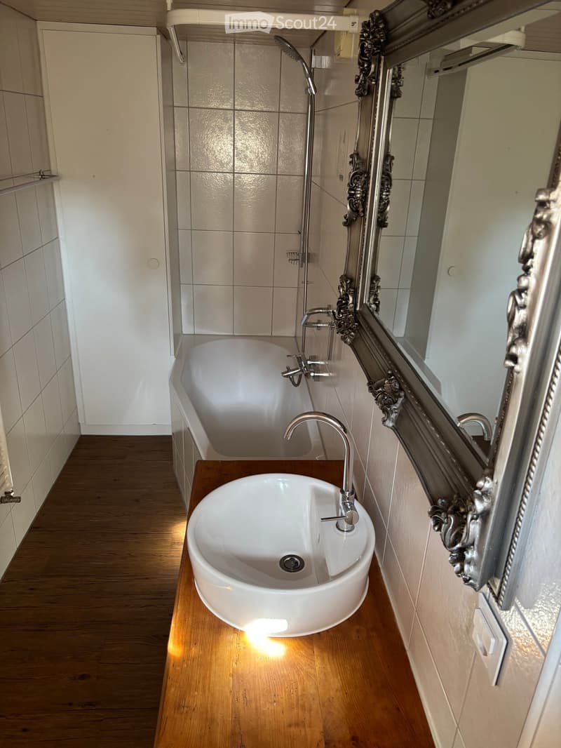 Modern bathroom with a bathtub, sink, and ornate mirror. The room features wooden flooring and tiled walls.