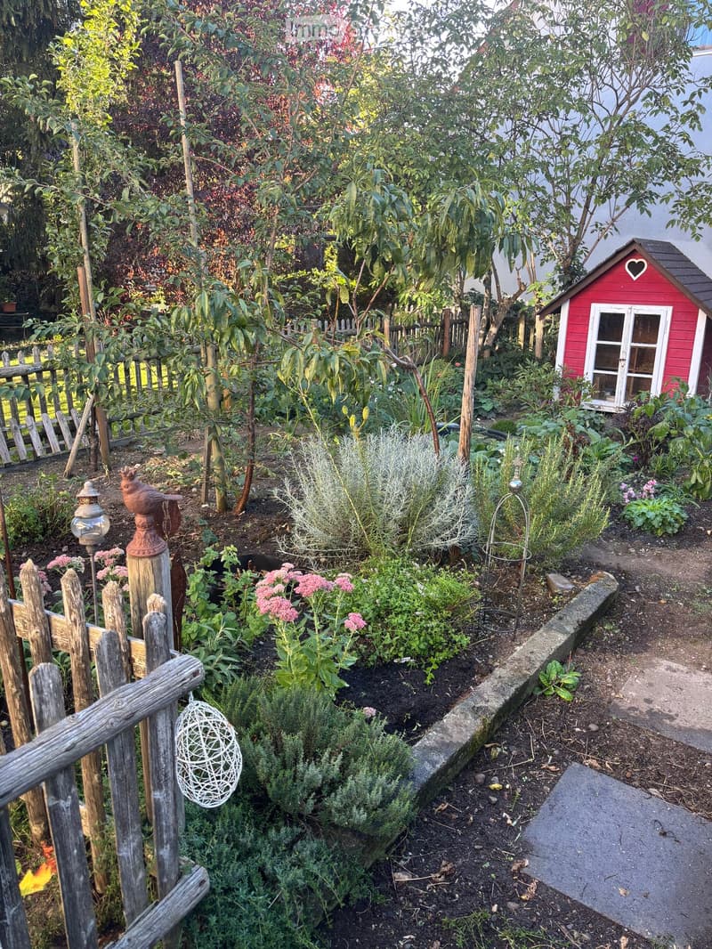 A well-maintained garden with a wooden fence, various plants, a small red shed, and decorative elements like a bird figurine and a hanging ornament.