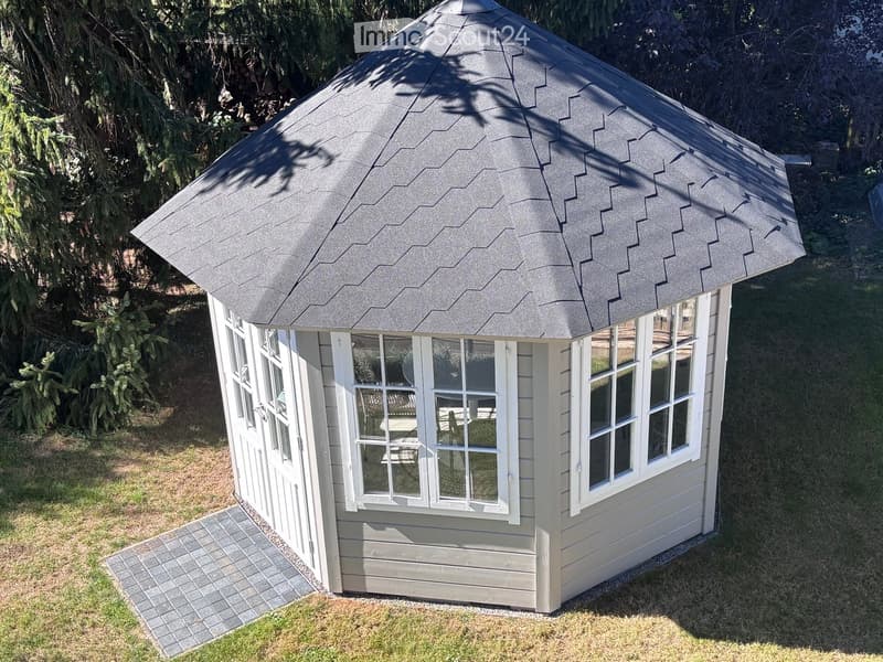 A small, octagonal wooden structure with a grey shingled roof and white-framed windows, situated on a paved pathway in a grassy area.