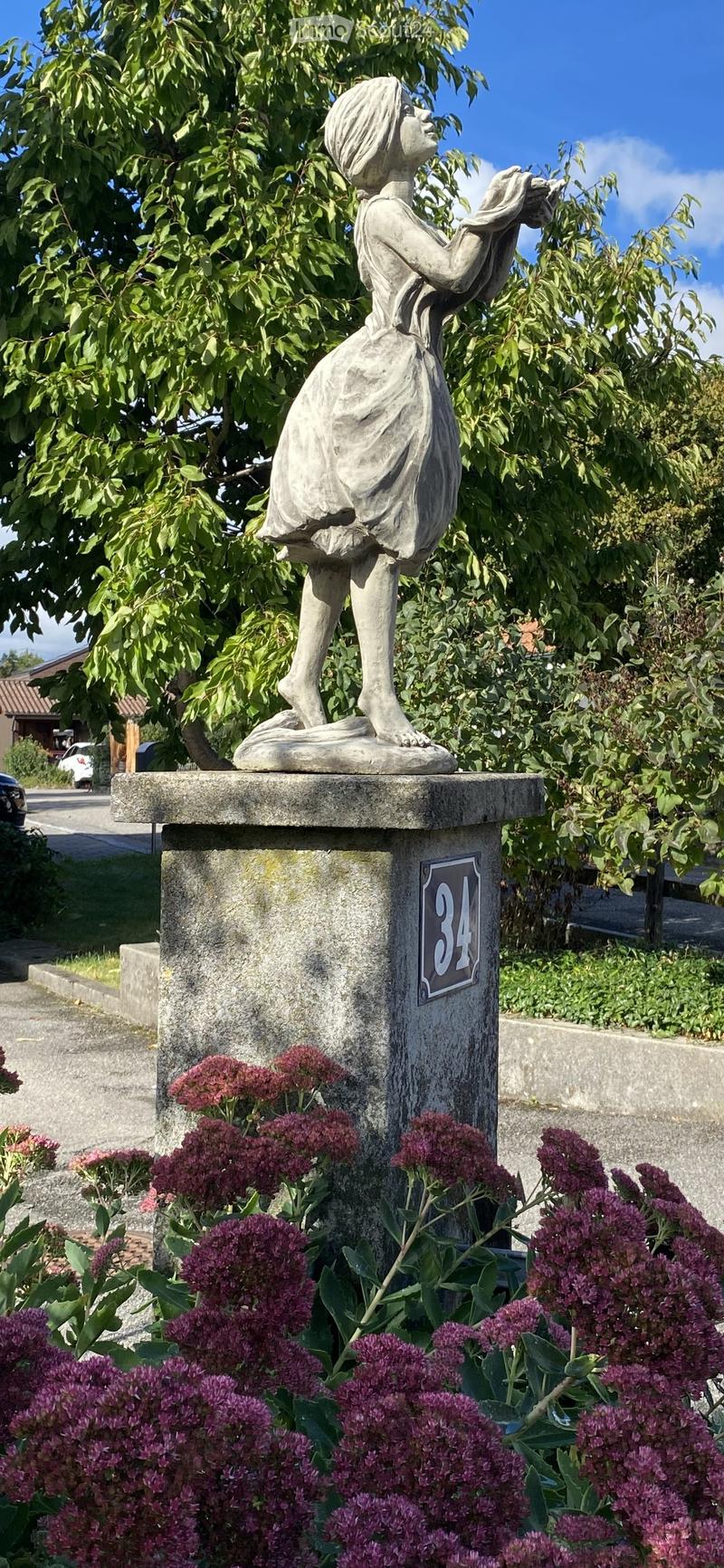 Garden area with a stone statue of a woman on a pedestal, surrounded by purple flowers and green foliage.