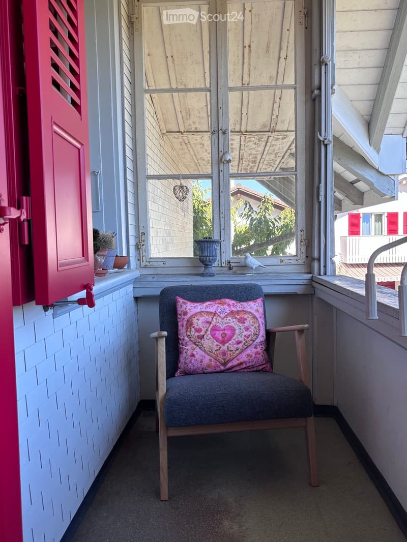 Balcony with a wooden chair, pink floral cushion, small vase, and a bird on the windowsill.