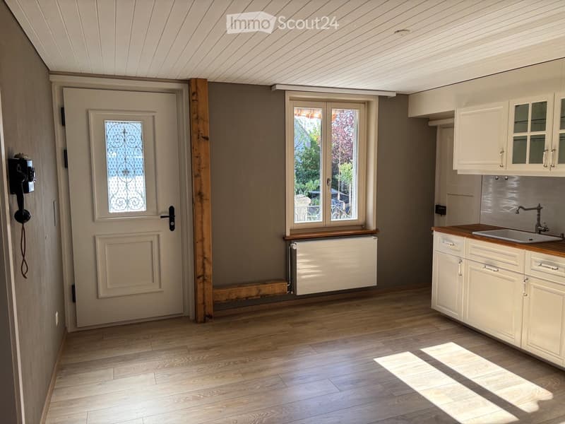 Empty room with wooden flooring, white cabinets, sink, and a door with decorative glass panel. Natural light streams in from a window.