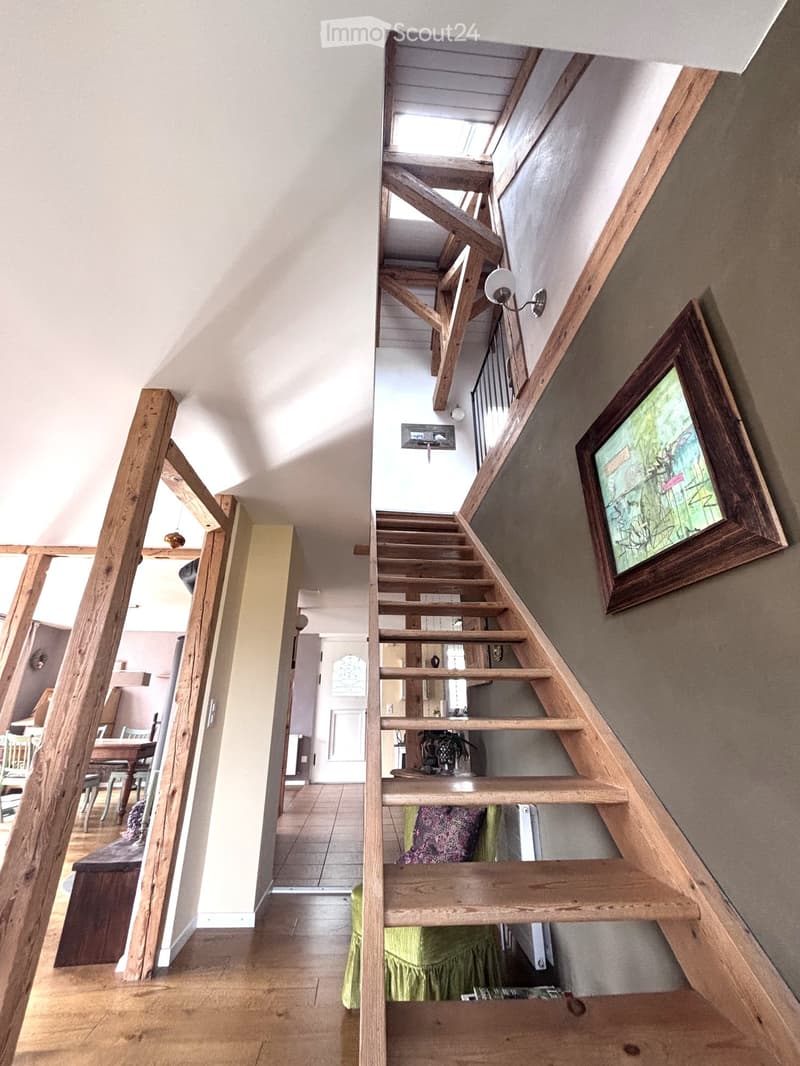 Wooden staircase with open wooden railings, leading to a room with visible furniture including a dining table and chairs, and a green upholstered chair in the foreground.