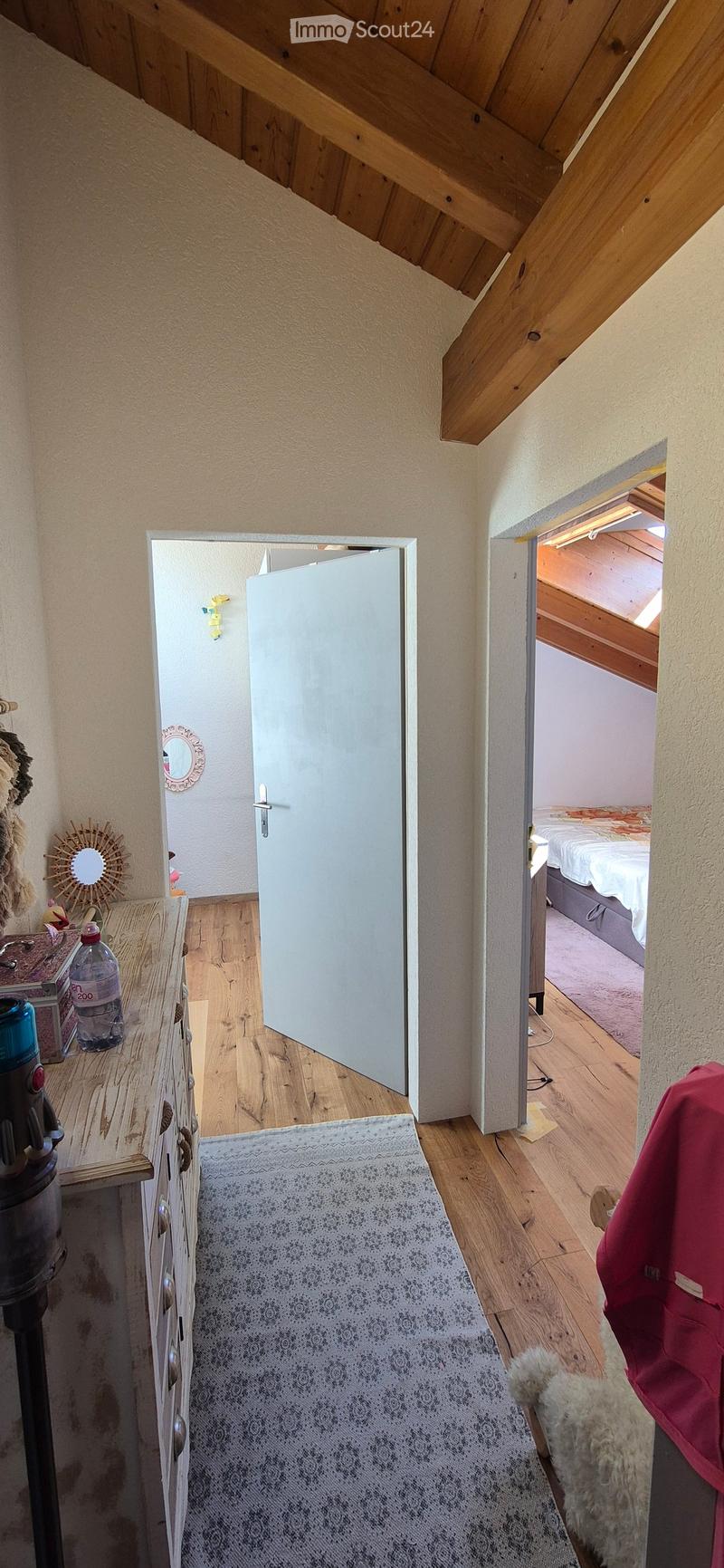 A narrow hallway with wooden flooring, leading to a bedroom. Visible items include a rug, a small table with a water bottle, and a mirror on the wall.
