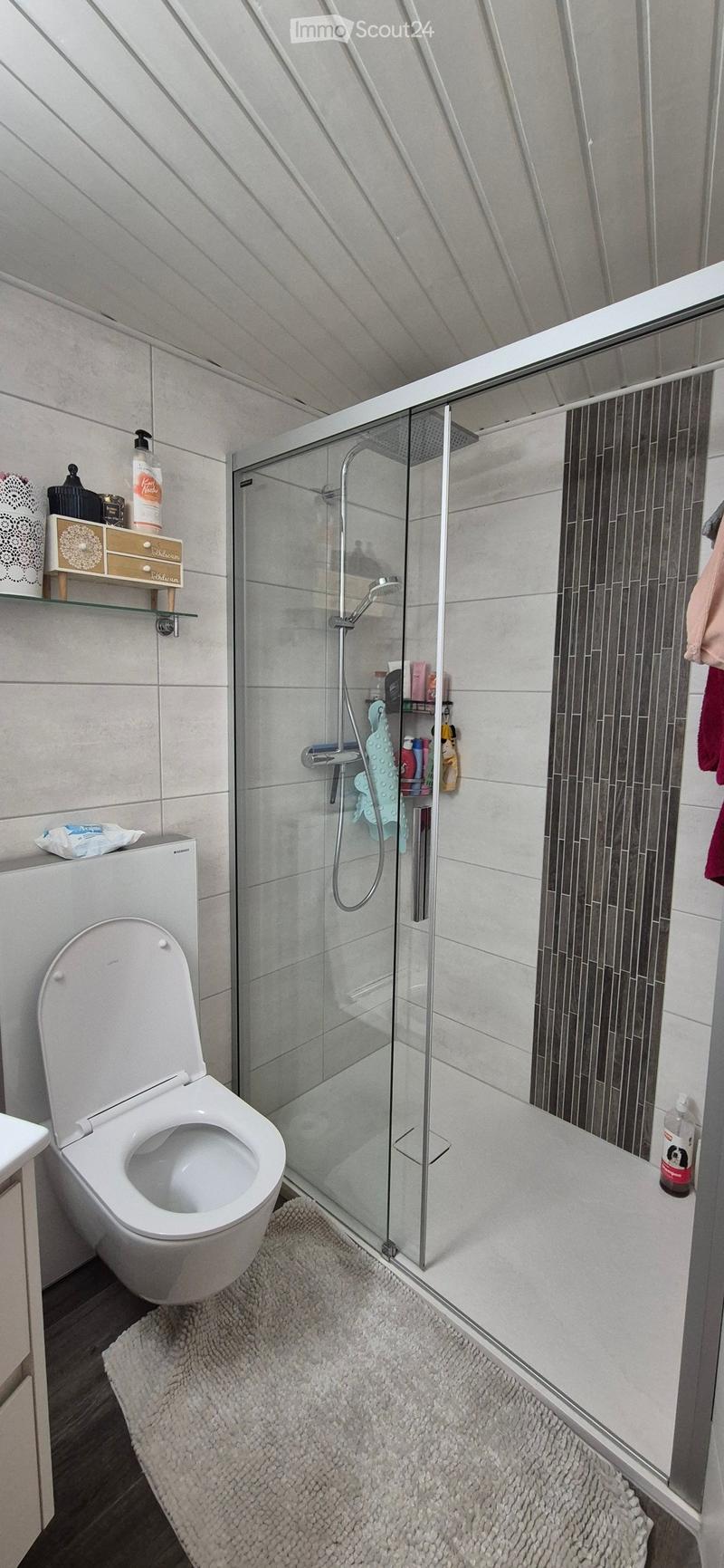 Modern bathroom with a toilet, glass-enclosed shower, and a small shelf holding toiletries.