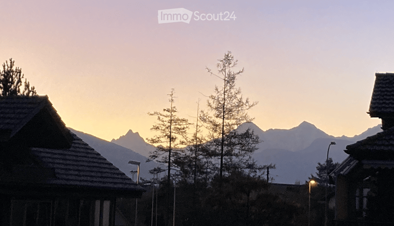 A serene outdoor scene featuring traditional wooden houses with sloped roofs, surrounded by slender trees and distant snow-capped mountains under a soft sunset sky.