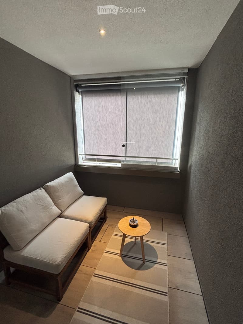 A small, cozy room with a corner sofa, a small round table, and a window with blinds.
