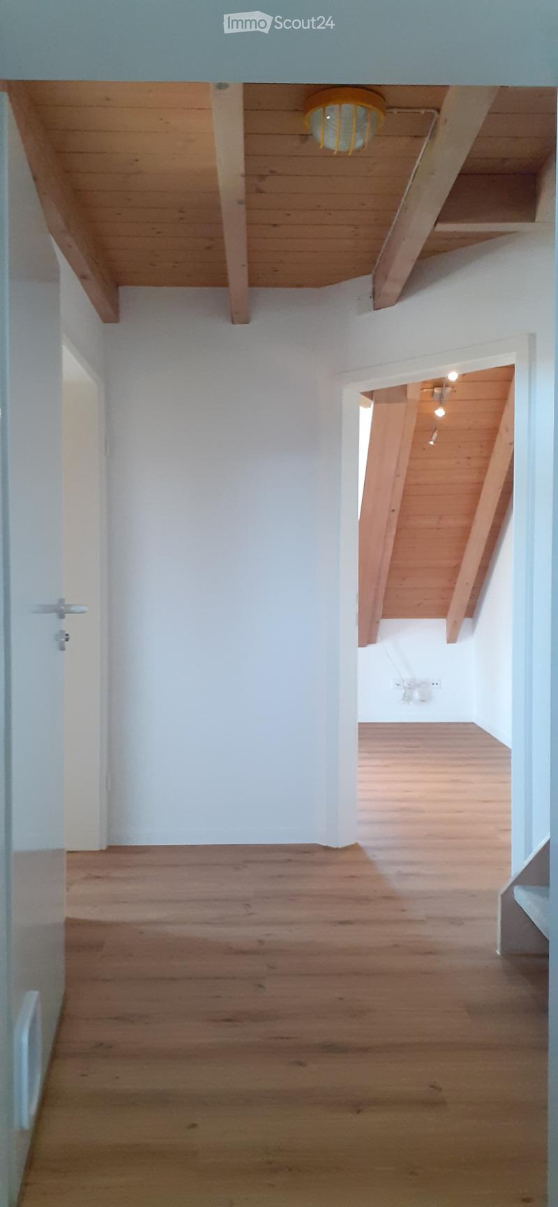 Empty hallway with wooden flooring, white walls, and exposed wooden beams on the ceiling. A doorway leads to another room with similar wooden flooring and a visible light fixture.