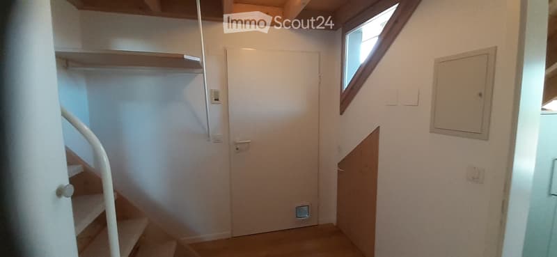A narrow hallway with a staircase leading upwards, a closed door, and a small window on the right side.