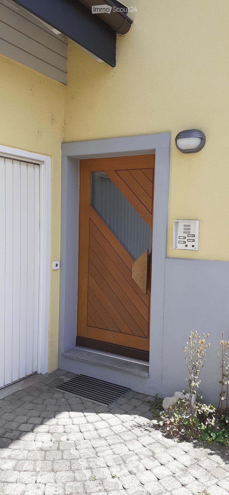 Exterior view of a building entrance with a wooden door, small steps, and a doormat. The building has a light yellow facade with a grey lower section.