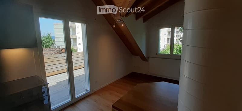 Bright, empty attic room with wooden flooring, large sliding glass door leading to a balcony, and exposed wooden beams on the ceiling.