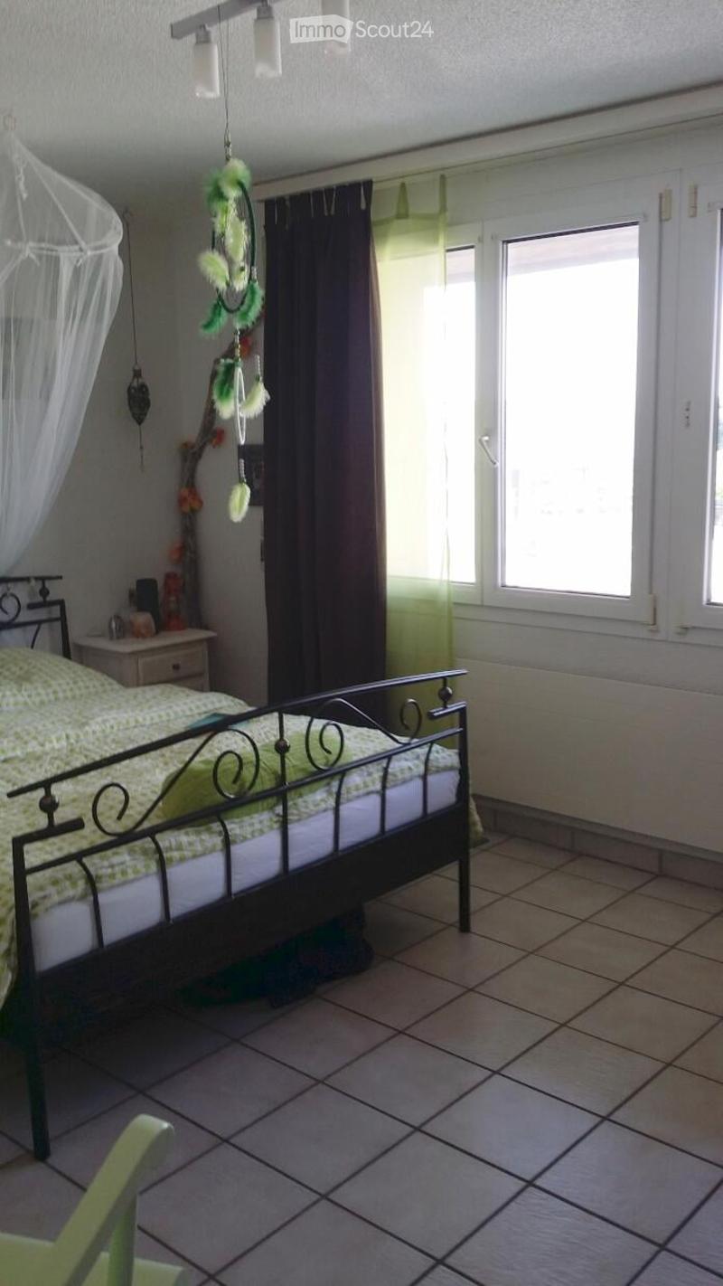 A cozy bedroom with a metal-framed bed, green bedding, a nightstand, a curtained window, and a dreamcatcher hanging from the ceiling.