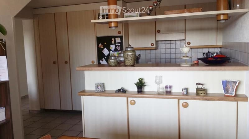 A modern kitchen with light wooden cabinets, a tiled backsplash, and a refrigerator. The countertops are adorned with various decorative items including jars, a teapot, a plant, and a glass vase.