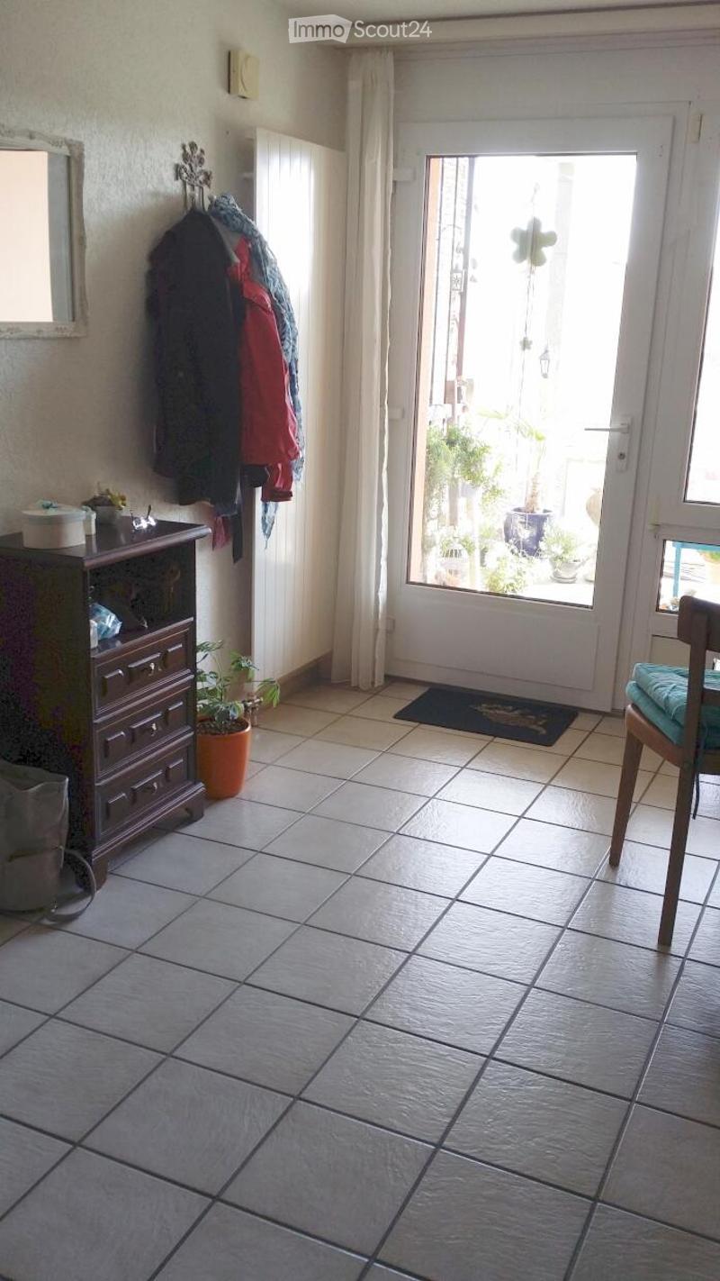 A small, well-lit hallway with tiled flooring, a coat rack with jackets, a small cabinet with drawers, a potted plant, a chair with a cushion, and a doormat near the entrance door leading to an outdoor area.