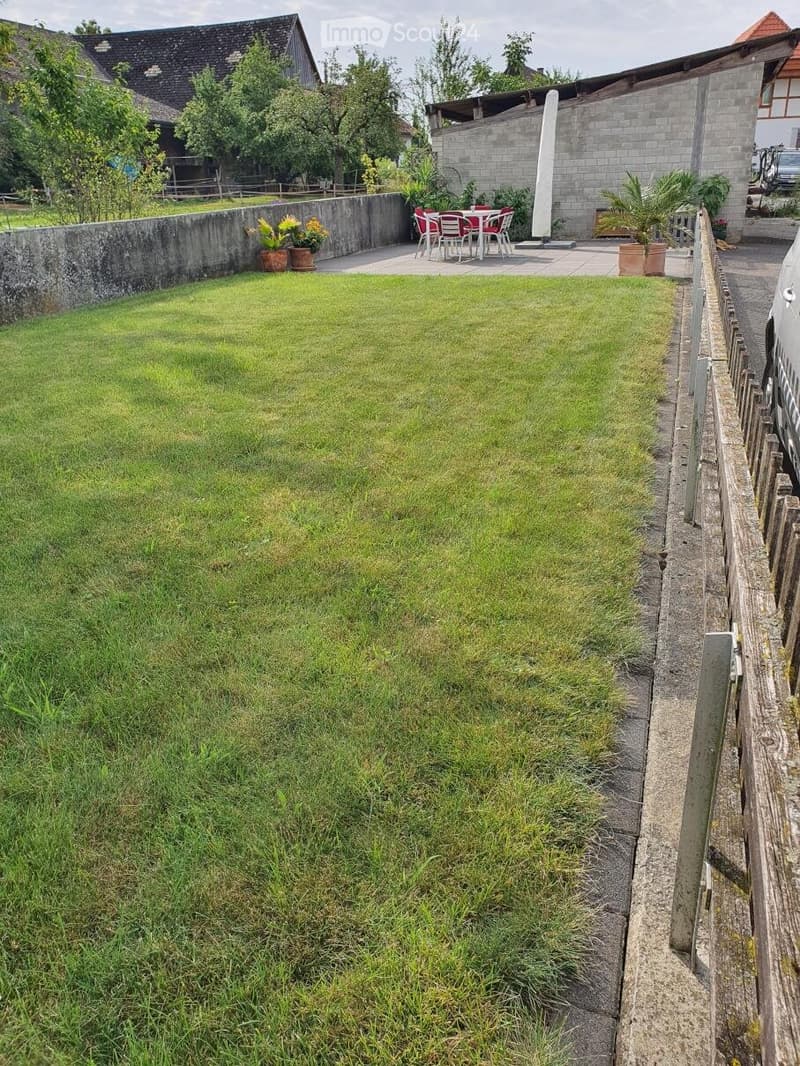 Well-maintained garden with a neatly trimmed lawn, surrounded by a low stone wall and wooden fence. There is a patio area with outdoor furniture, including a table and chairs, and potted plants.