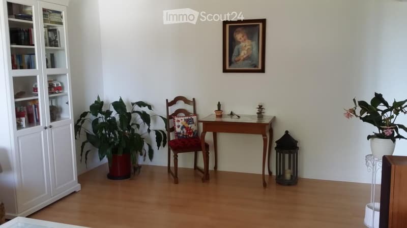 A cozy, well-lit room with wooden flooring, a wooden chair with a colorful cushion, a small wooden table with a plant and decorative items, a white bookcase with books and toys, and a framed picture on the wall.