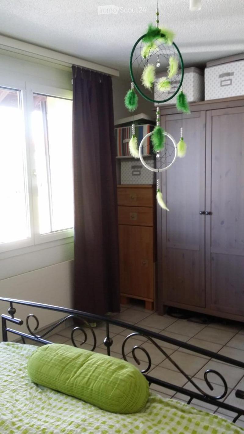 A cozy bedroom with a green and white patterned bed, a dreamcatcher hanging from the ceiling, and a wooden cabinet with books and storage boxes.
