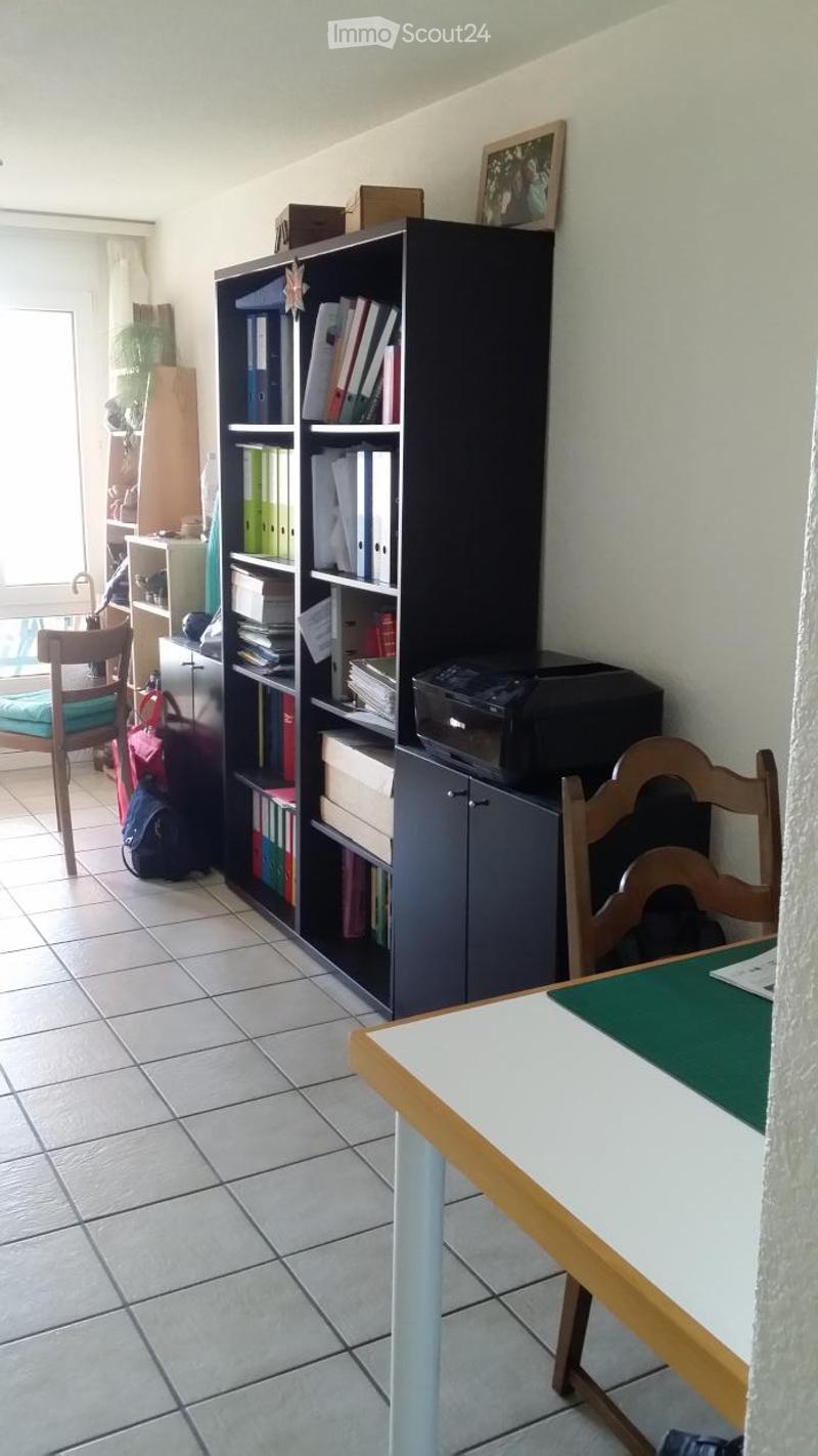 A narrow hallway with tiled walls, a wooden railing, and a large open shelving unit filled with books and storage boxes. The floor is light-colored, and there is a small framed picture on the floor near the shelving unit.