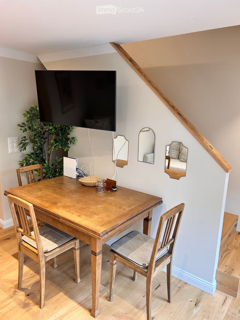 A small, cozy room with a wooden table and chairs, a mounted TV screen, a plant, and a staircase leading upwards.