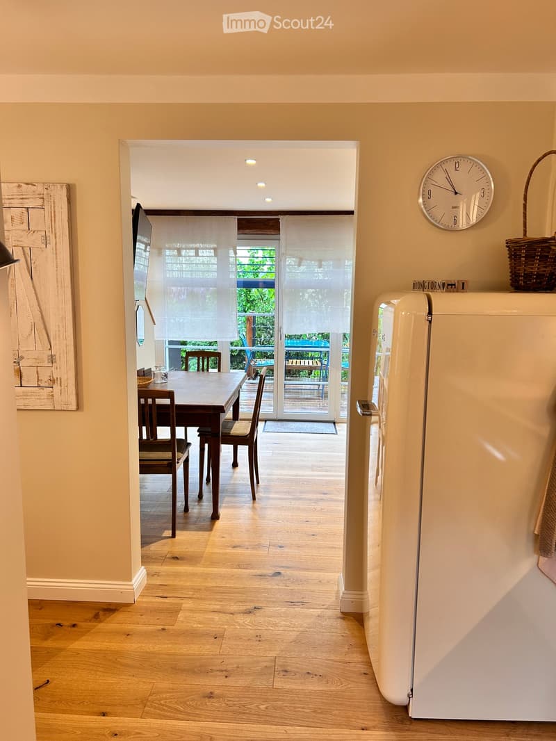 A well-lit hallway with wooden flooring leads to a dining area featuring a wooden table and chairs, a white refrigerator, and a wall-mounted clock. A woven basket sits on top of the refrigerator, and a doorway opens to a balcony with outdoor furniture vis