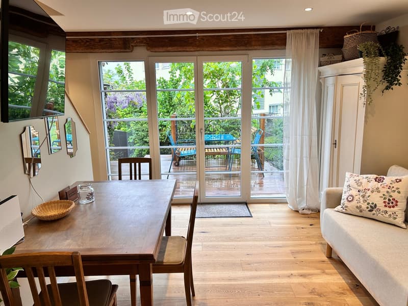 Bright living and dining area with wooden floors, large windows, and sliding glass doors leading to a balcony. Features a sofa, dining table with chairs, and decorative mirrors.
