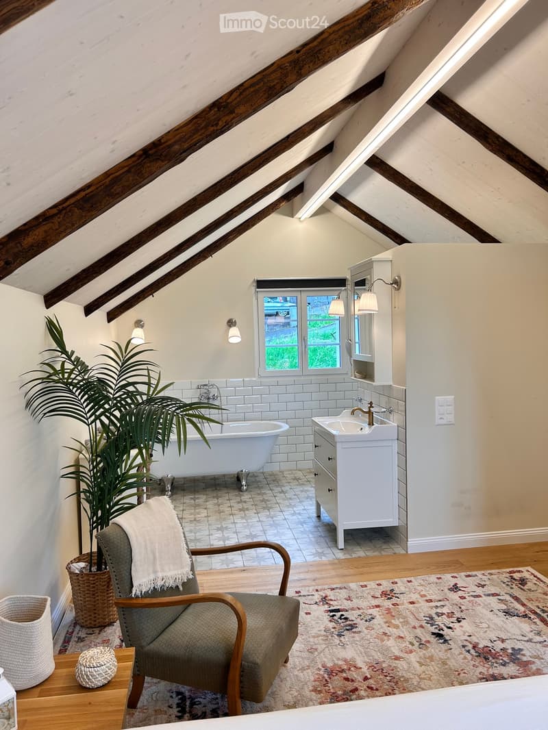 Bathroom with exposed wooden beams, a freestanding bathtub, a vanity with double sinks, a chair with a blanket, and a patterned rug.