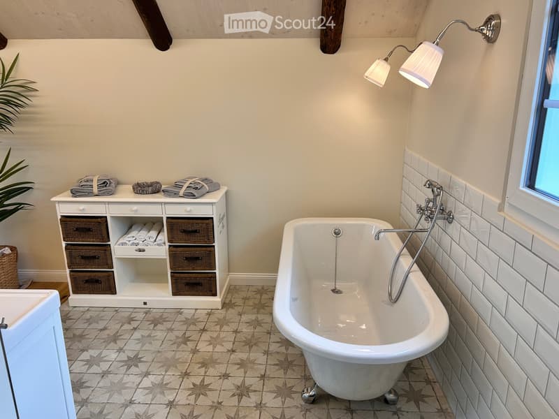 Bathroom with a white clawfoot bathtub, white tiled wall, storage cabinet with wicker baskets, folded towels, and a wall-mounted lamp.