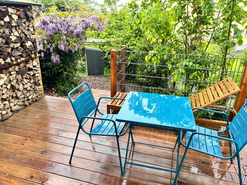 Wooden deck with a blue metal table and two matching chairs, surrounded by lush greenery and a wooden fence.