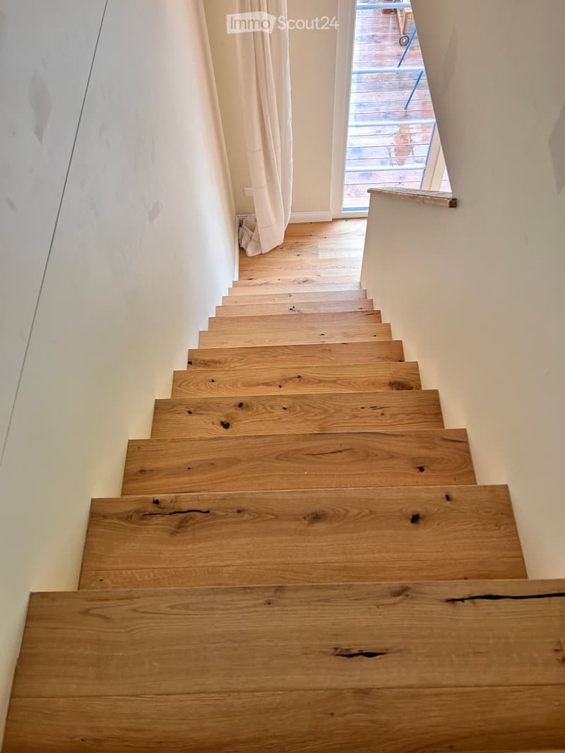 Wooden staircase with light-colored steps and railings, leading to an open area with a view of a wooden deck outside.