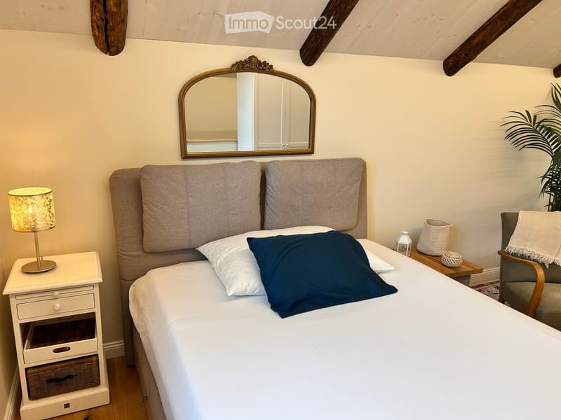 Cozy bedroom with a large bed, nightstand with lamp, decorative mirror, and a small wooden table with a wicker basket and lantern.