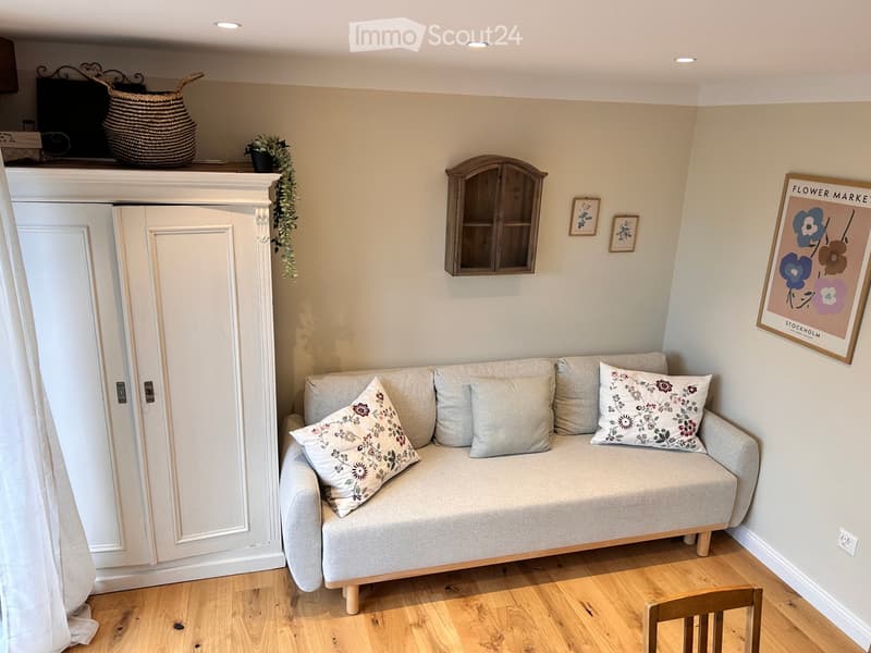 Cozy living room with a light grey sofa adorned with floral pillows, a small wooden cabinet, framed artwork, a woven basket, and a potted plant on the mantelpiece.