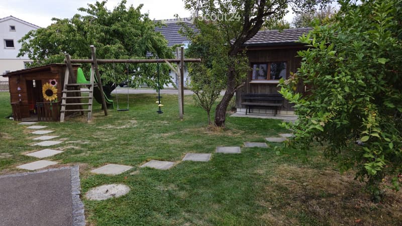 A well-maintained garden with a wooden playhouse, swing set, slide, and a bench. The area is surrounded by trees and has a stone pathway.