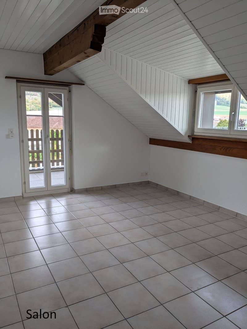 Empty attic room with slanted wooden ceiling, tiled floor, large window, and sliding glass door leading to a balcony.