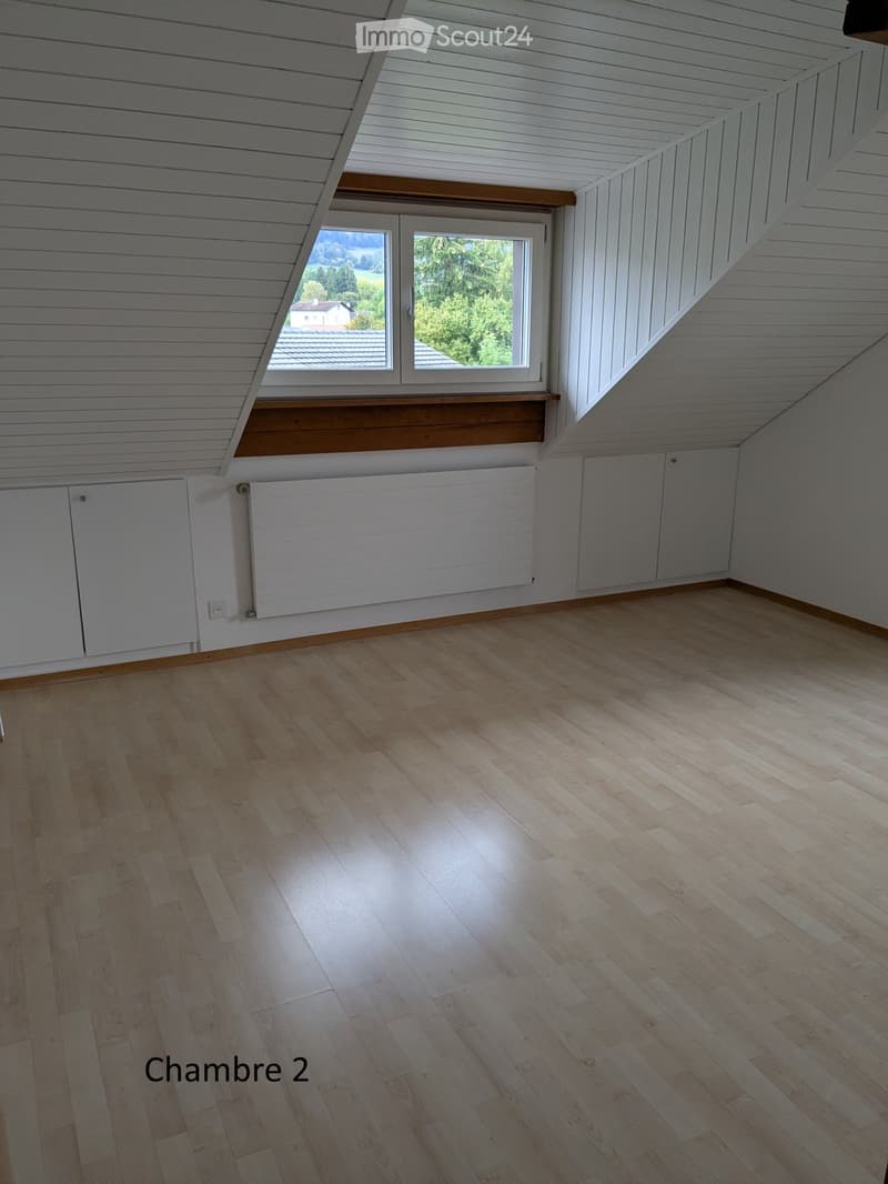 Empty attic room with wooden flooring, white paneled walls, and a large window offering a view of the outdoors.