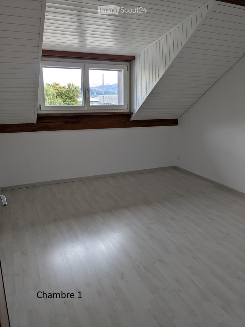 Empty room with light wooden flooring, white walls, and a large window with white frames. The ceiling has white paneling with wooden trim around the window area.